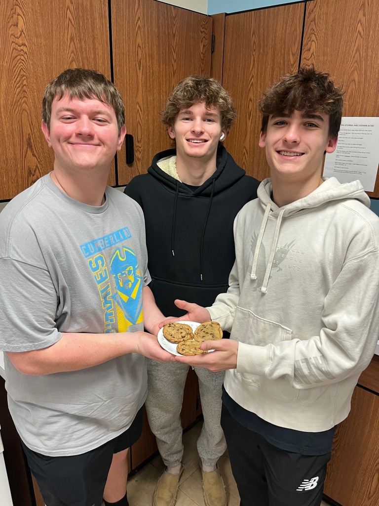 Three students are standing side by side. They are holding a plate of three cookies. 