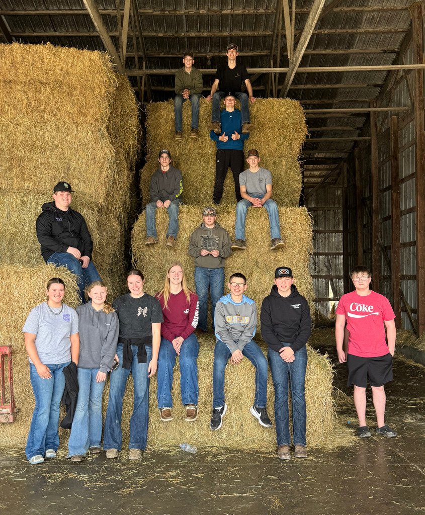 The 14 members of the team are standing against, sitting on, and standing on bales of hay. 