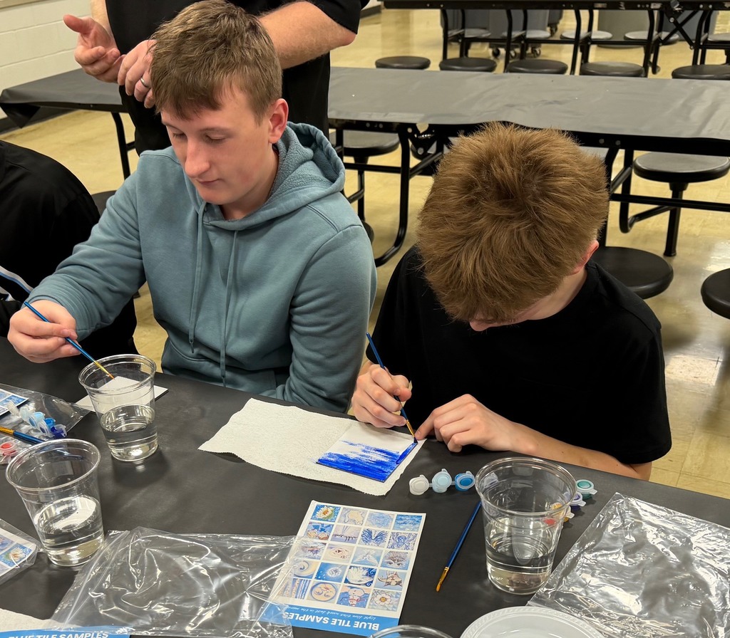 Students are sitting at a table painting a  white tile with blue paint. 