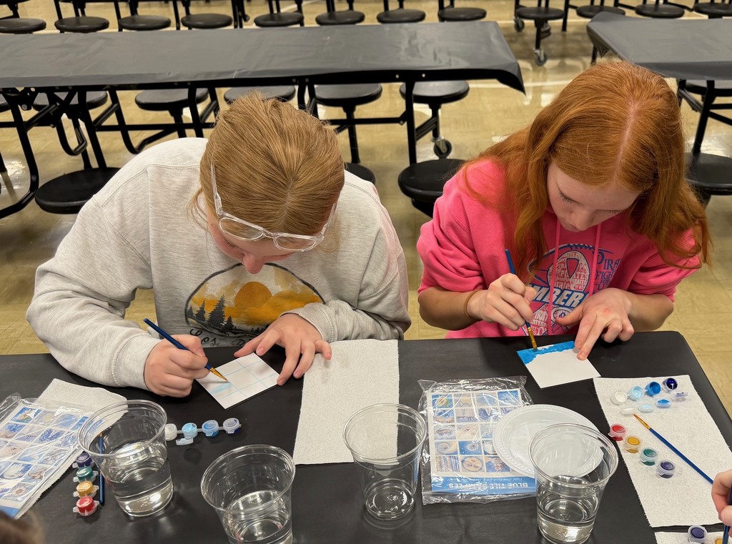 Two students are sitting at a table painting a  white tile with blue paint. 