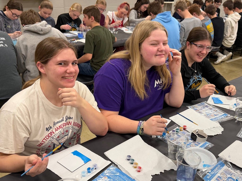Three students are sitting at a table painting a  white tile with blue paint. 