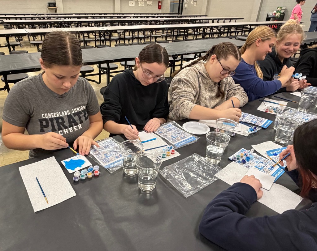 Six students are sitting at a table painting a  white tile with blue paint. 