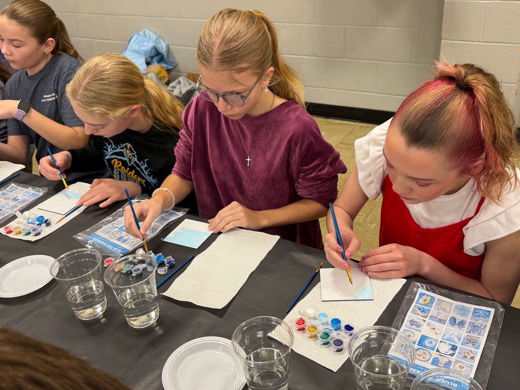 Three students are sitting at a table painting a  white tile with blue paint. 