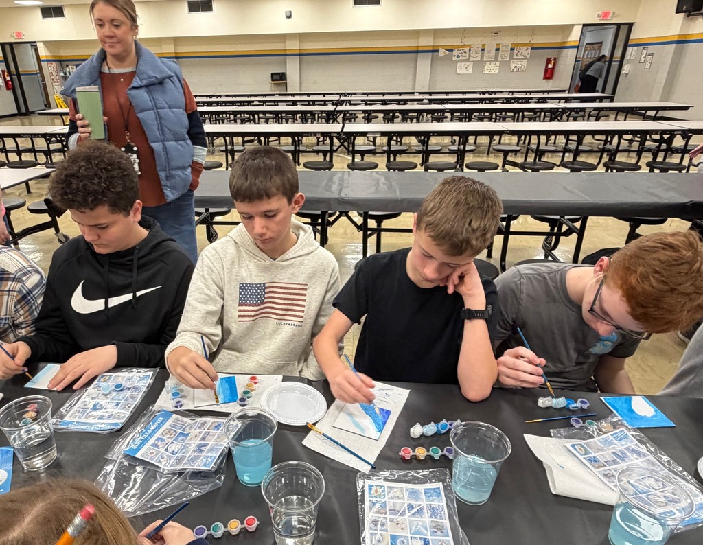 Four students are sitting at a table painting a  white tile with blue paint. 