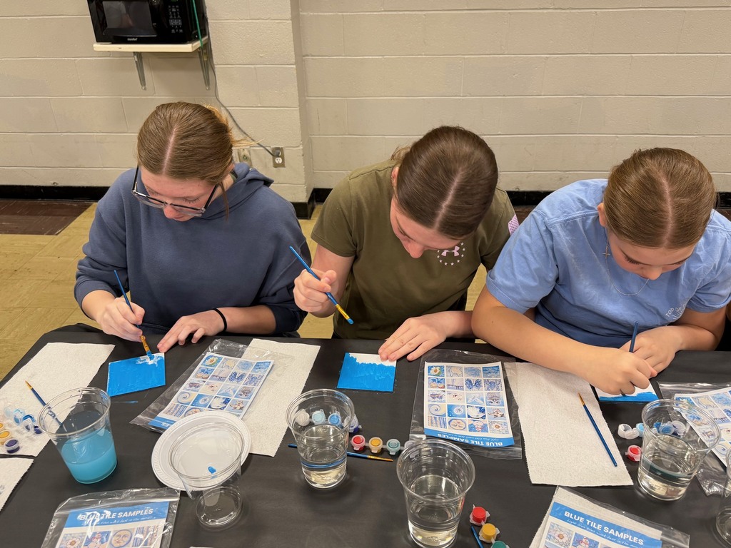 Three students are sitting at a table painting a  white tile with blue paint. 