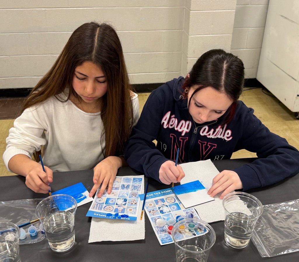 Two students are sitting at a table painting a  white tile with blue paint. 