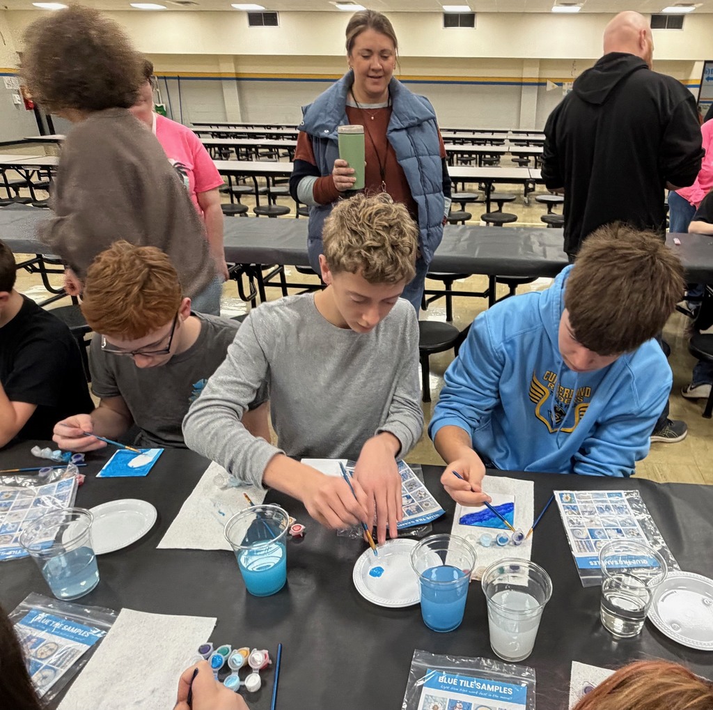 Three students are sitting at a table painting a  white tile with blue paint. 