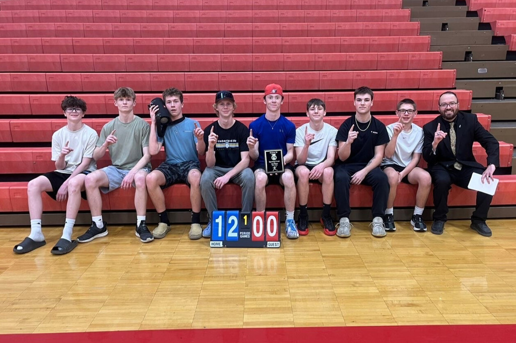 The team of eight is sitting on the bleachers with their teacher. They are holding the 1st place plaque. 