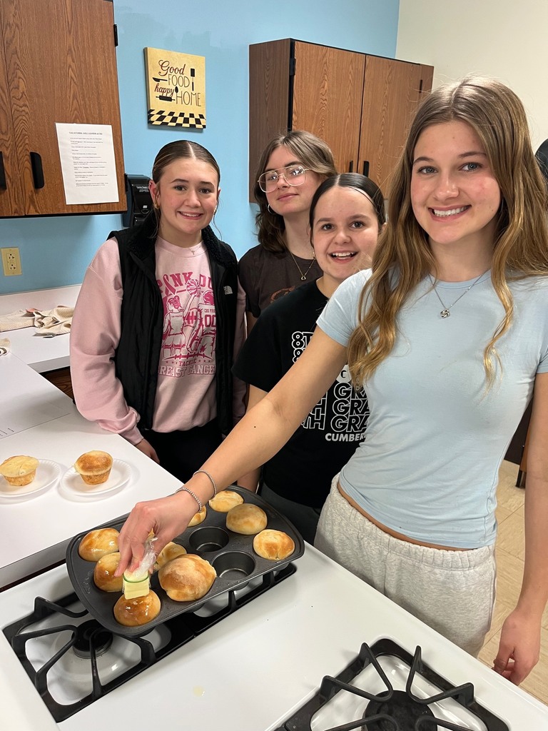 Four students are standing by their pan of baked rolls. One is buttering the rolls. 