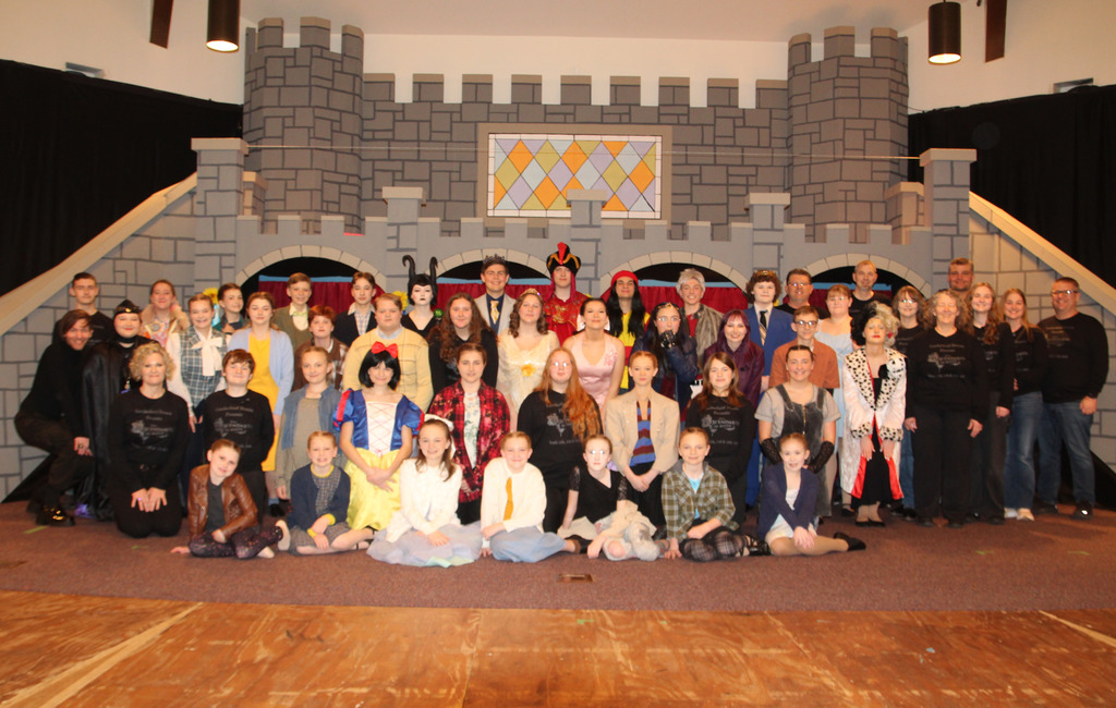 The cast and crew of Descendants the Musical are sitting or standing in one of four rows. They are in costume in front of a castle background.
