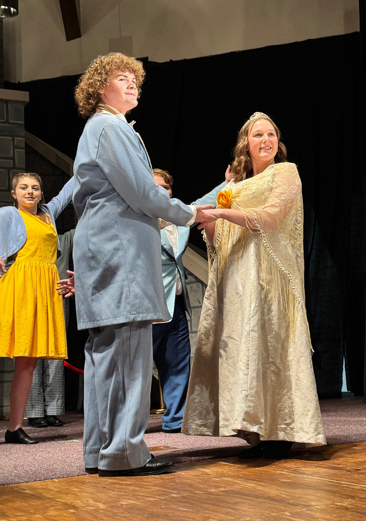Two cast members, one in a gold dress and another in a suit, are dancing. The cast is behind dancing as well.