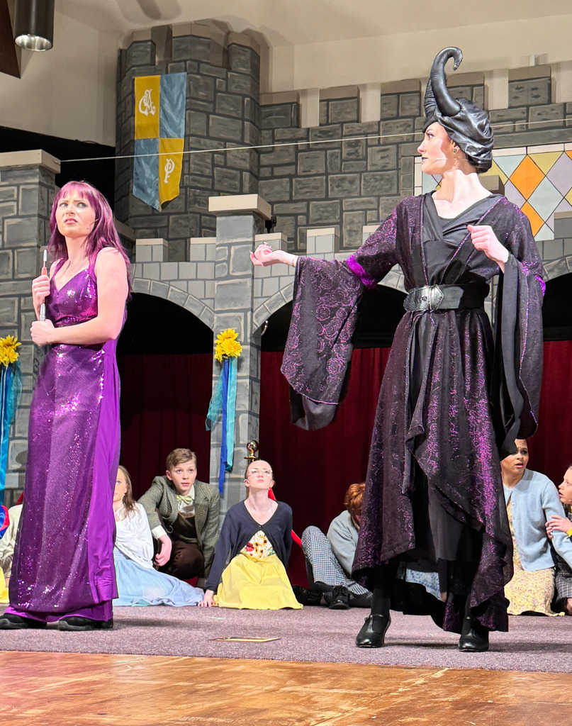 Two characters from the musical, both dressed in purple, are taking to each other. THere are other cast members sitting on the ground in the background.
