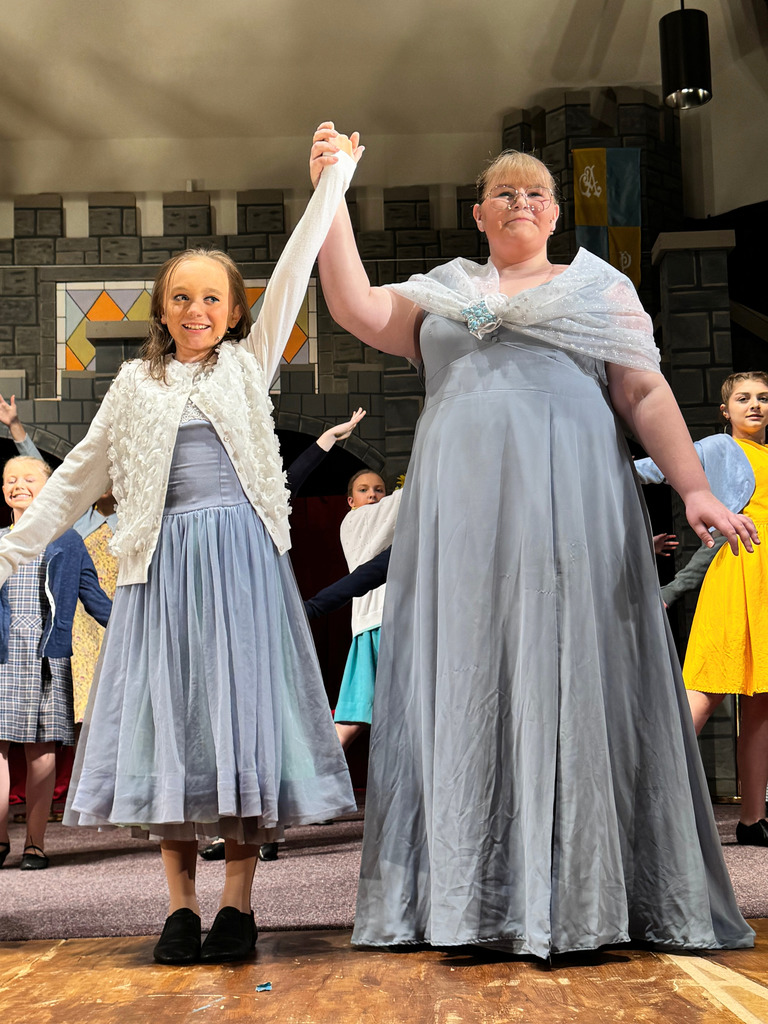 Two cast members, both in blue dresses, are dancing. The cast is behind dancing as well.