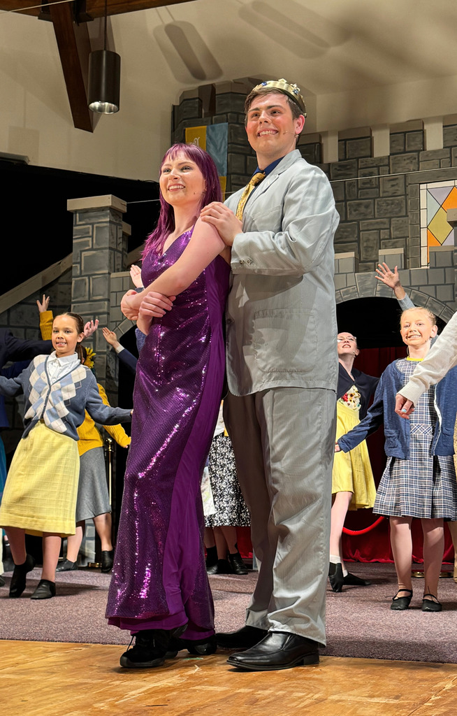 Two cast members, one in a gold dress and another in a suit, are dancing. The cast is behind dancing as well.