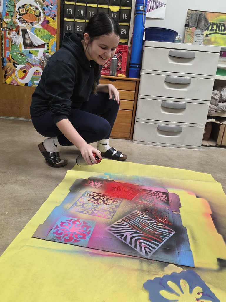 A student is squatting on the ground and using spraypaint to create designs on the cardboard piece.