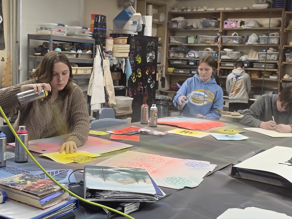 Two students are standing at a table and using spraypaint to create designs on their cardboard piece.