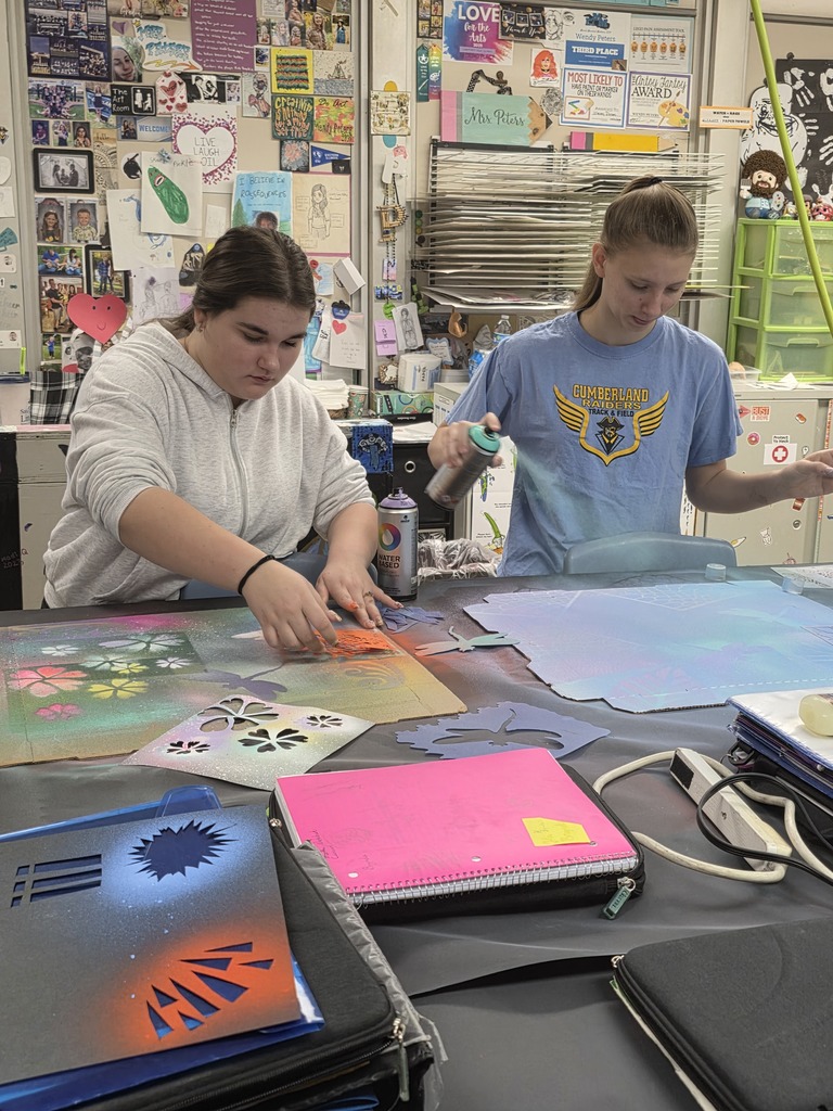 Two students are standing at a table and using spraypaint to create designs on their cardboard piece.