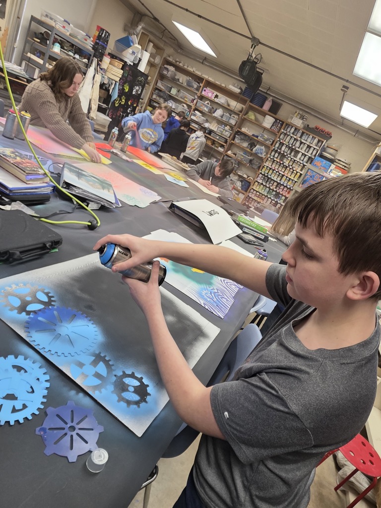 A student is standing at a table and using spraypaint to create designs on the cardboard piece.