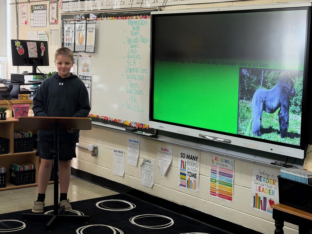 A student is standing at the podium sharing the student's slide show that is presented on the whiteboard.