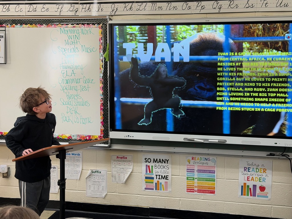 A student is standing at the podium sharing the student's slide show that is presented on the whiteboard.