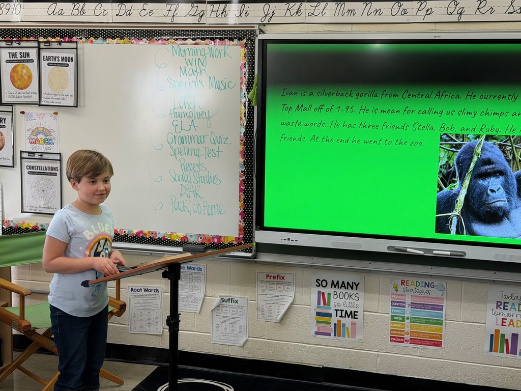 A student is standing at the podium sharing the student's slide show that is presented on the whiteboard.