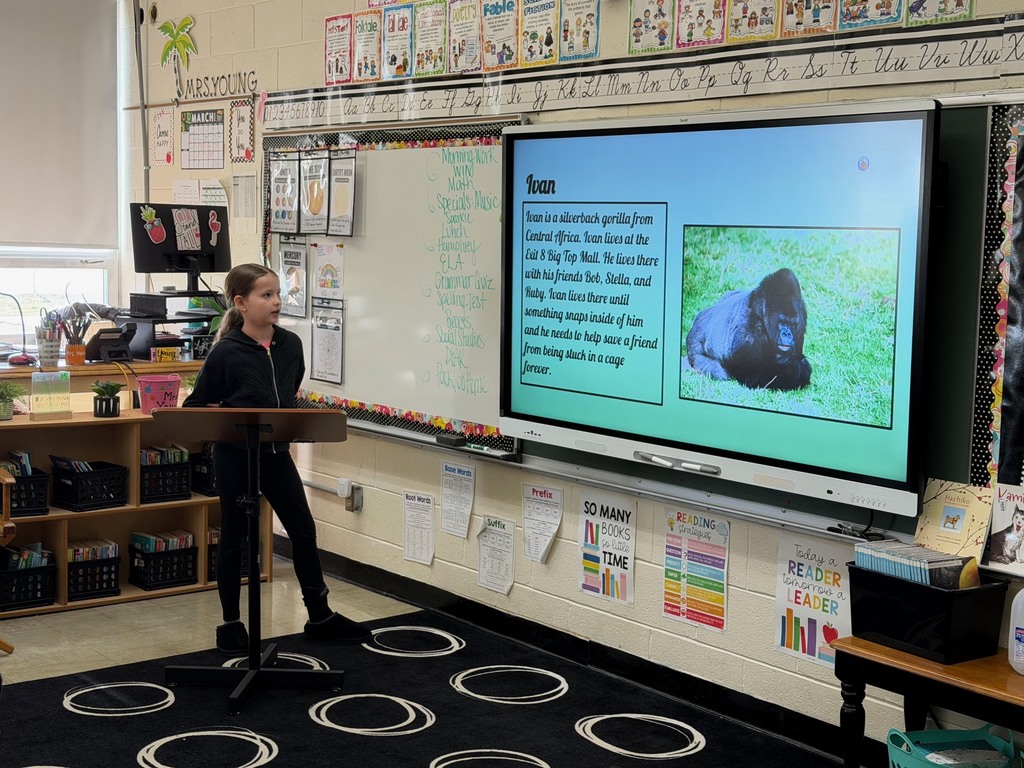 A student is standing at the podium sharing the student's slide show that is presented on the whiteboard.