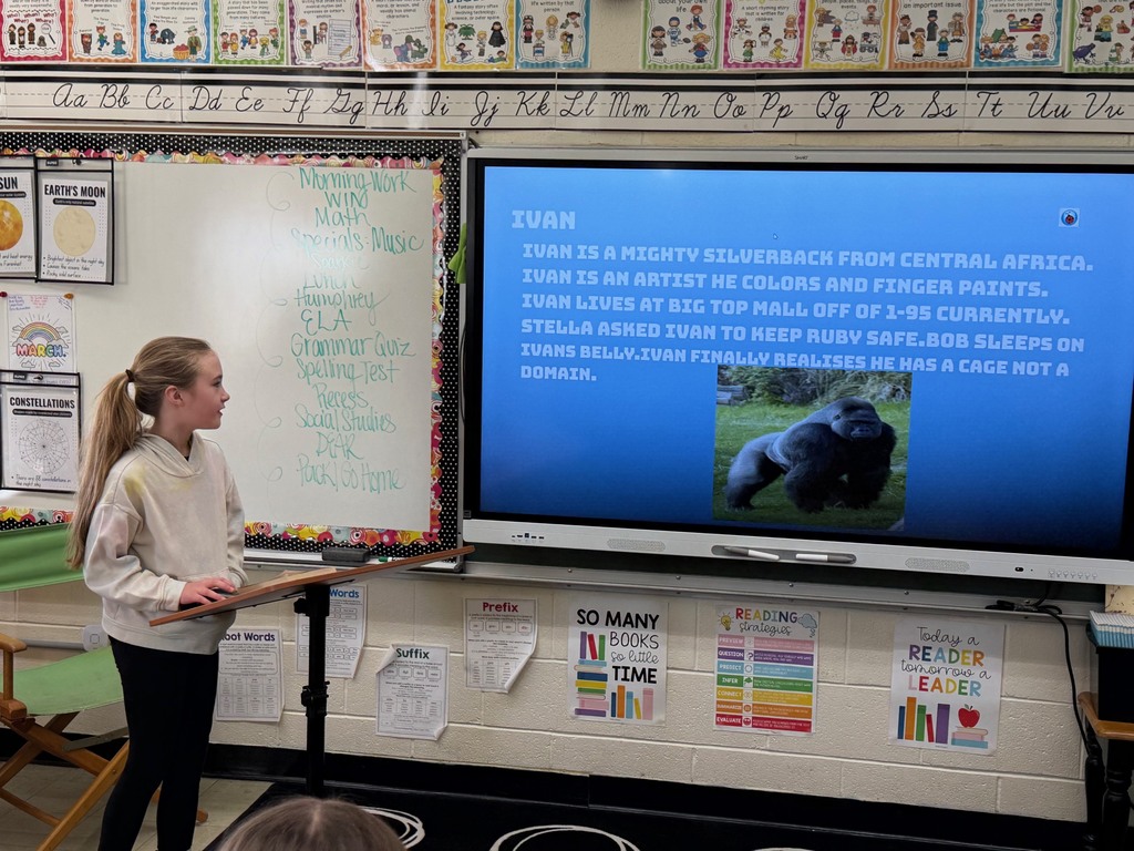 A student is standing at the podium sharing the student's slide show that is presented on the whiteboard.