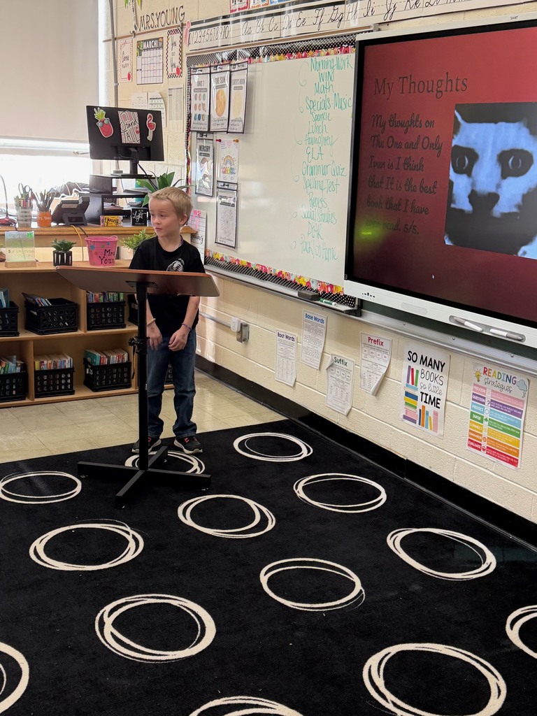 A student is standing at the podium sharing the student's slide show that is presented on the whiteboard.