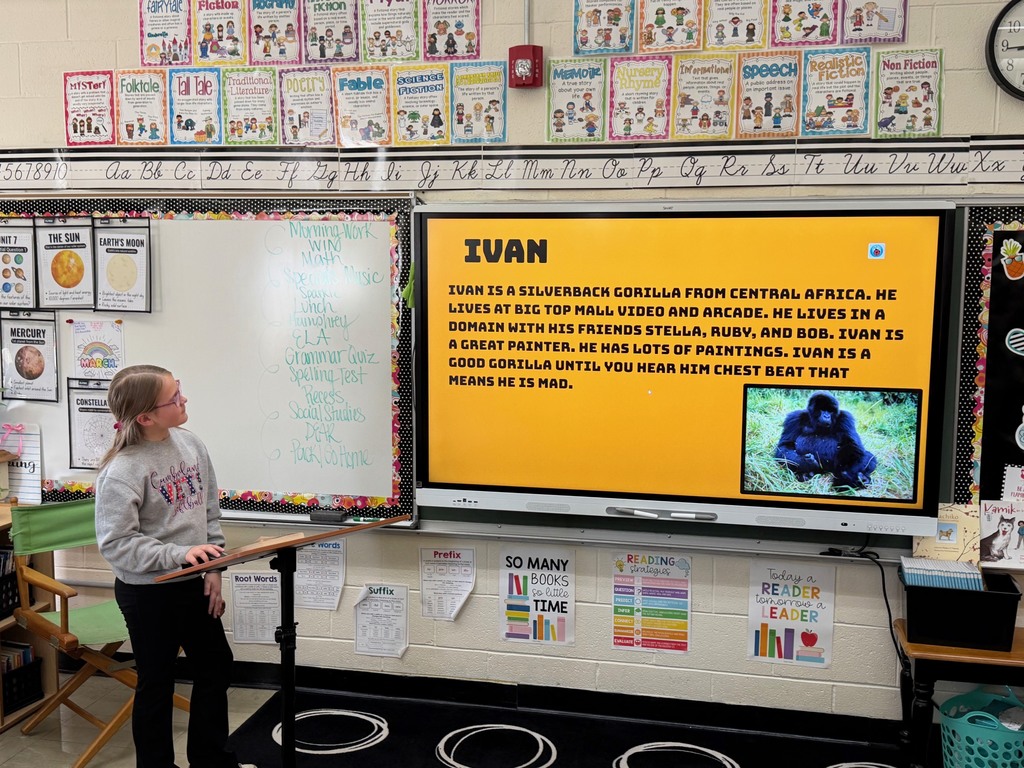 A student is standing at the podium sharing the student's slide show that is presented on the whiteboard.