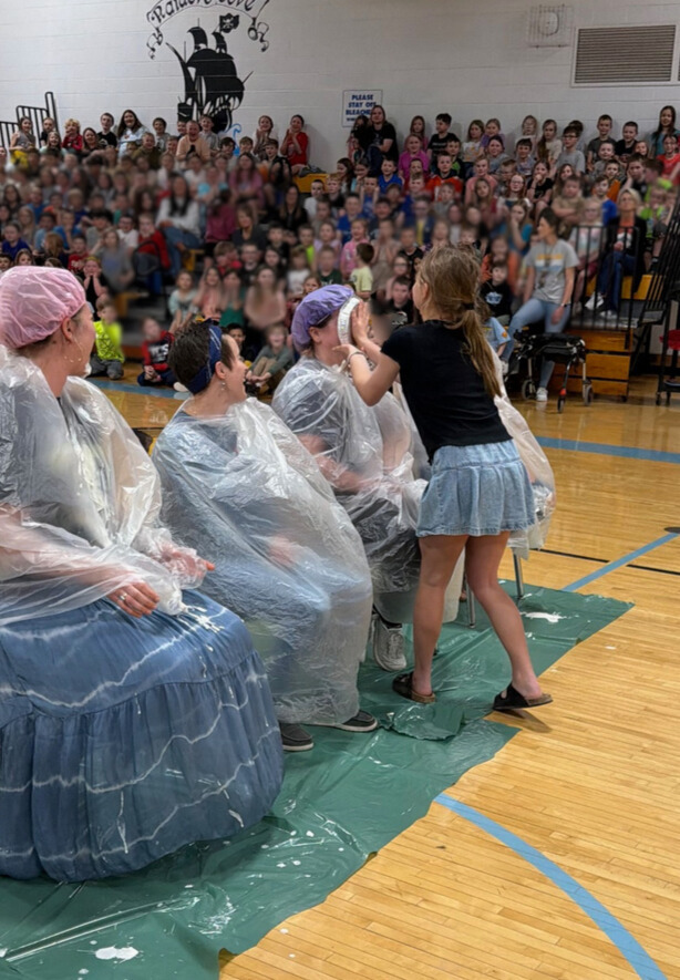 A student is throwing a pie in the face of a staff member. Staff members are sitting with trash bags on in the middle of the basketball court. 
