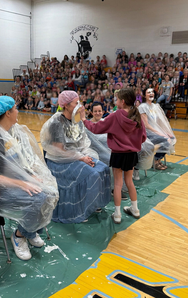 A student is throwing a pie in the face of a staff member. Staff members are sitting with trash bags on in the middle of the basketball court. 