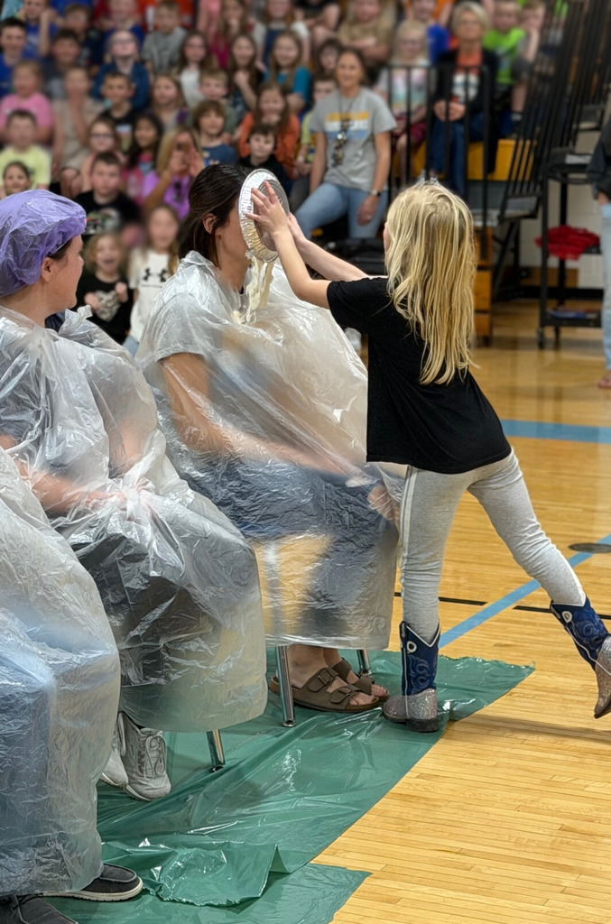 A student is throwing a pie in the face of a staff member. Staff members are sitting with trash bags on in the middle of the basketball court. 