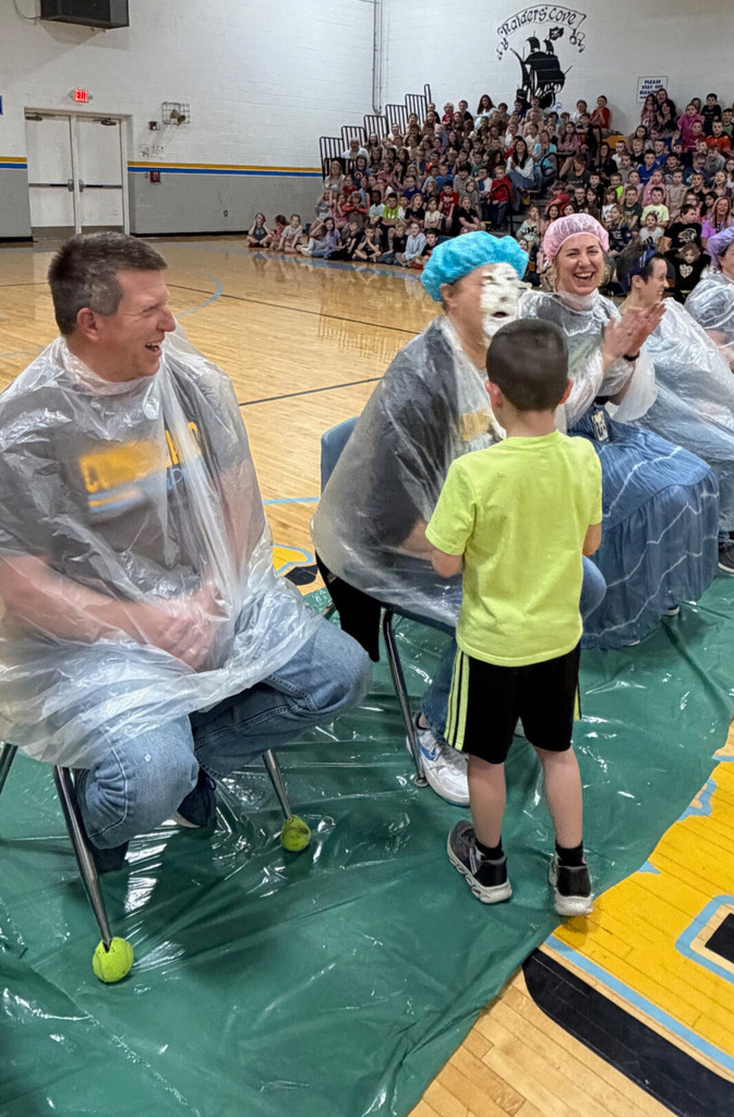 A student is throwing a pie in the face of a staff member. Staff members are sitting with trash bags on in the middle of the basketball court. 