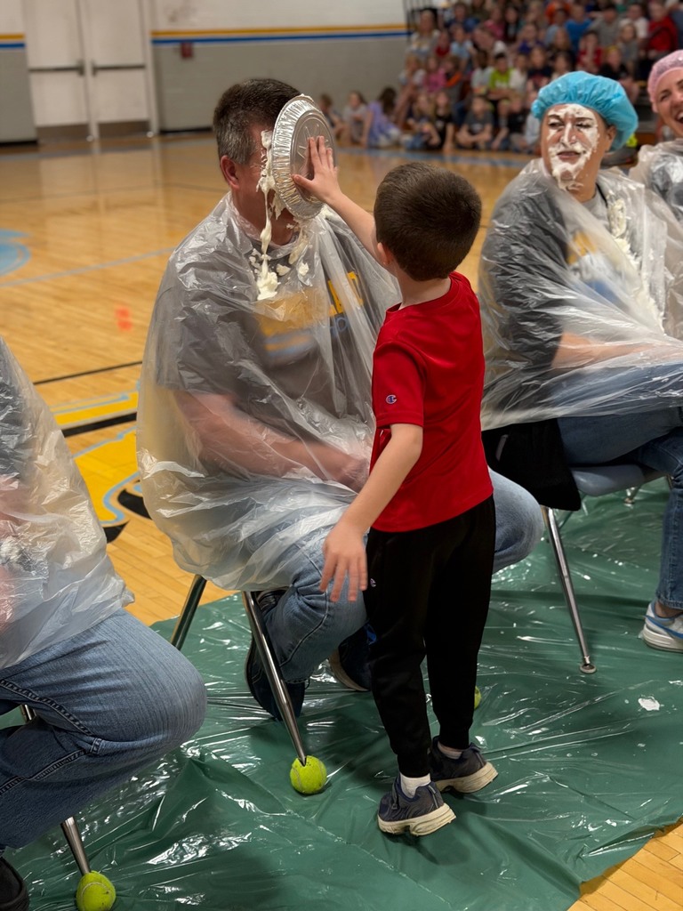 A student is throwing a pie in the face of a staff member. Staff members are sitting with trash bags on in the middle of the basketball court. 