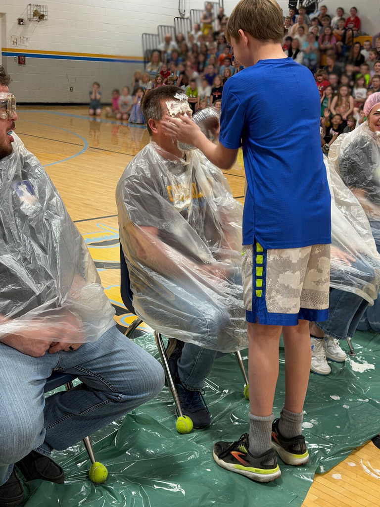 A student is throwing a pie in the face of a staff member. Staff members are sitting with trash bags on in the middle of the basketball court. 