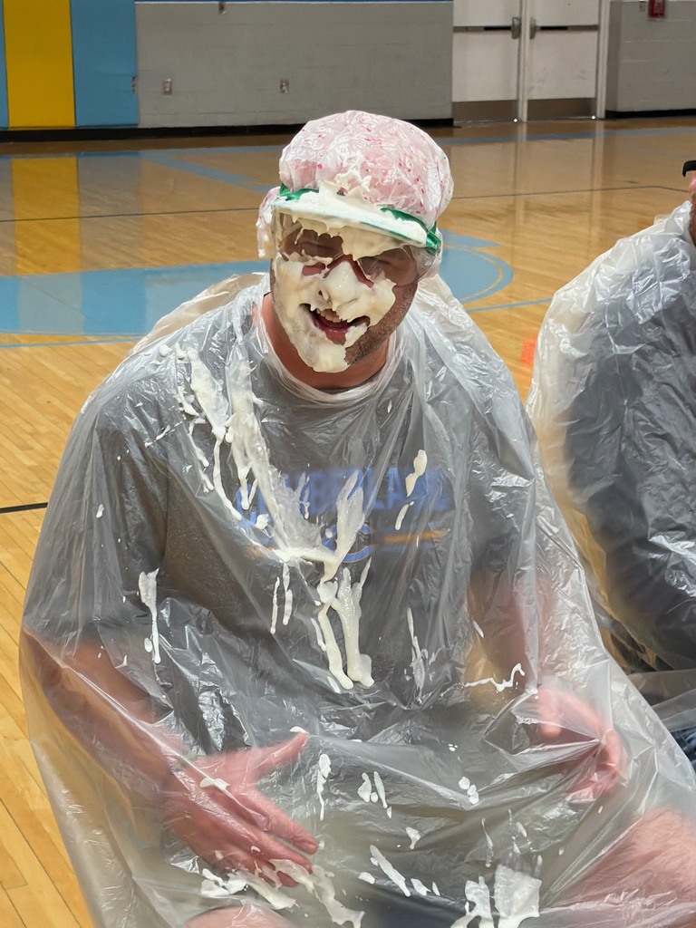 A staff member is pictured just after being pied.  Staff members are sitting with trash bags on in the middle of the basketball court. 
