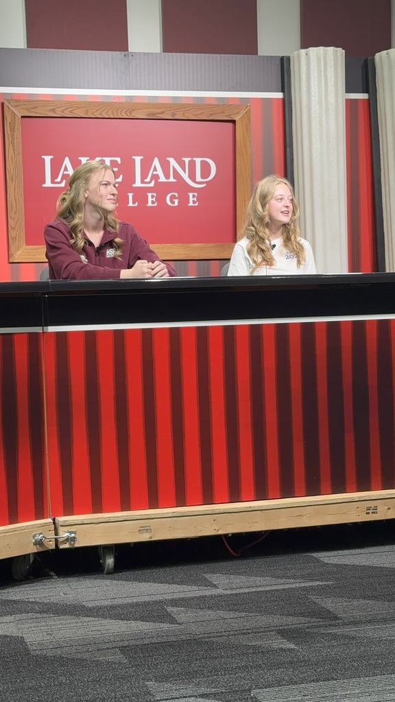 Two students are sitting at the desk for a news broadcast. 