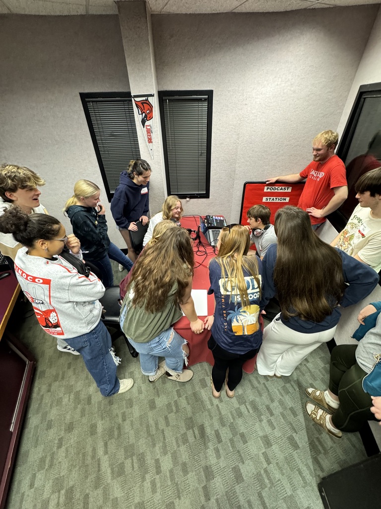 A group of 13 students are standing around a table listing to a radio broadcast. 