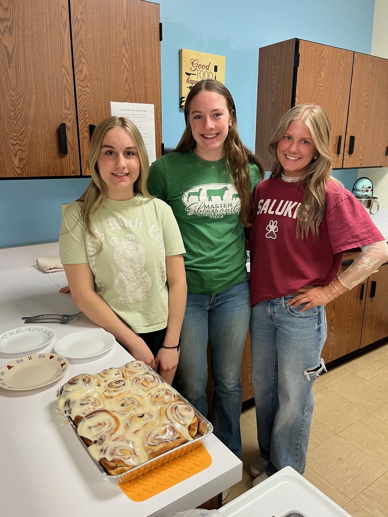 Three students are standing by the counter by their pan of cinnamon rolls. 
