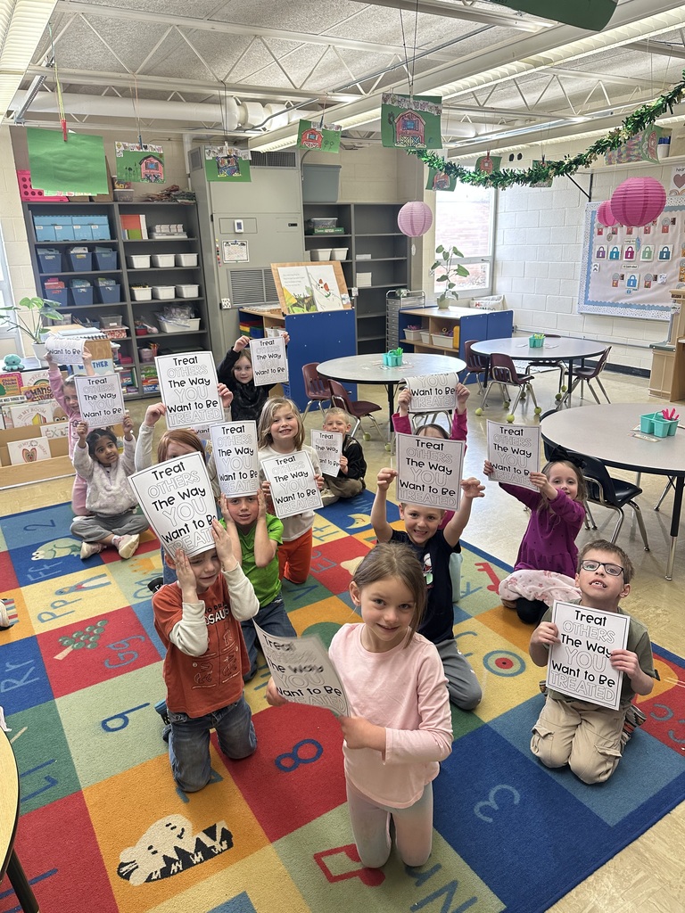 The students are sitting on the carpet. Each is holding a paper that reads Treat others the way You want to be teated.
