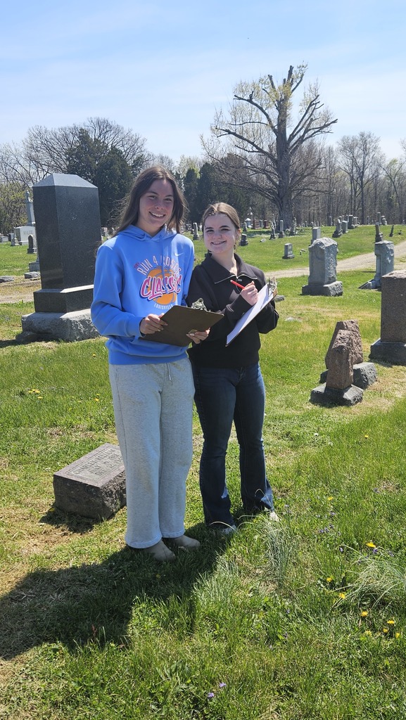 Two students are standing near a gravestone with a clipboard collecting data.