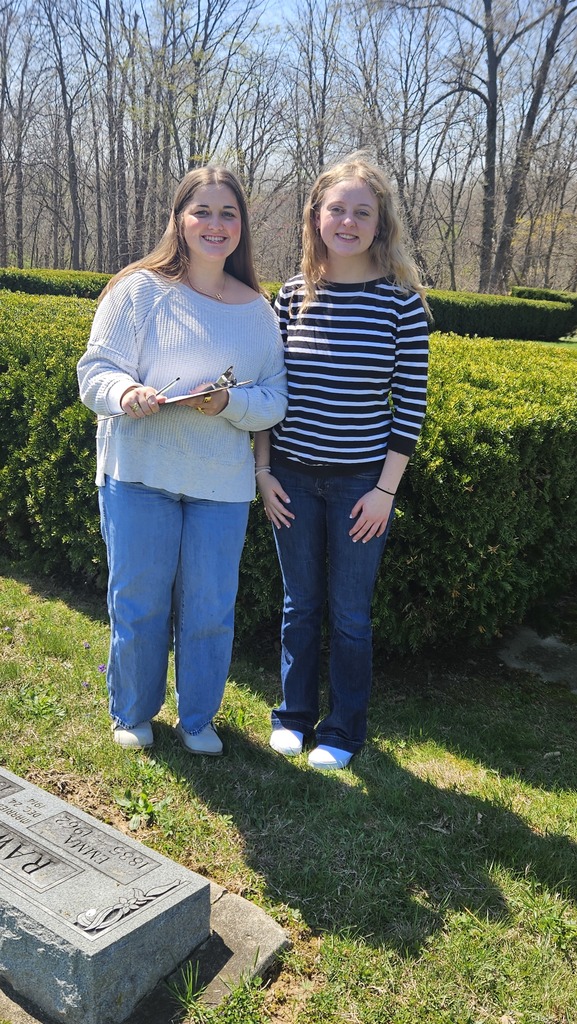 Two students are standing near a gravestone with a clipboard collecting data.