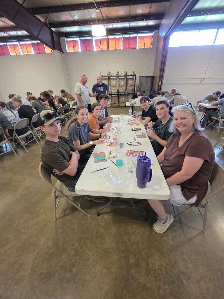 A large group of students are sitting at the tables painting their tiles.