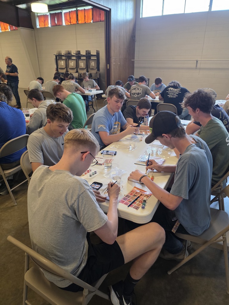 A large group of students are sitting at the tables painting their tiles.