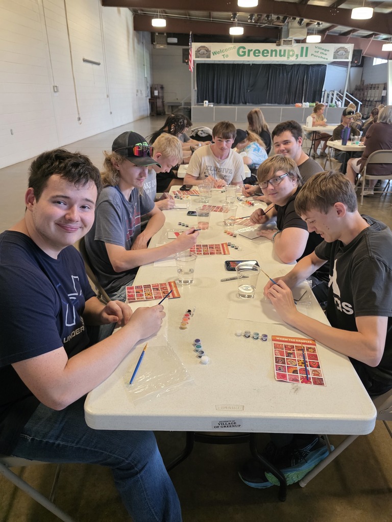 A large group of students are sitting at the tables painting their tiles.