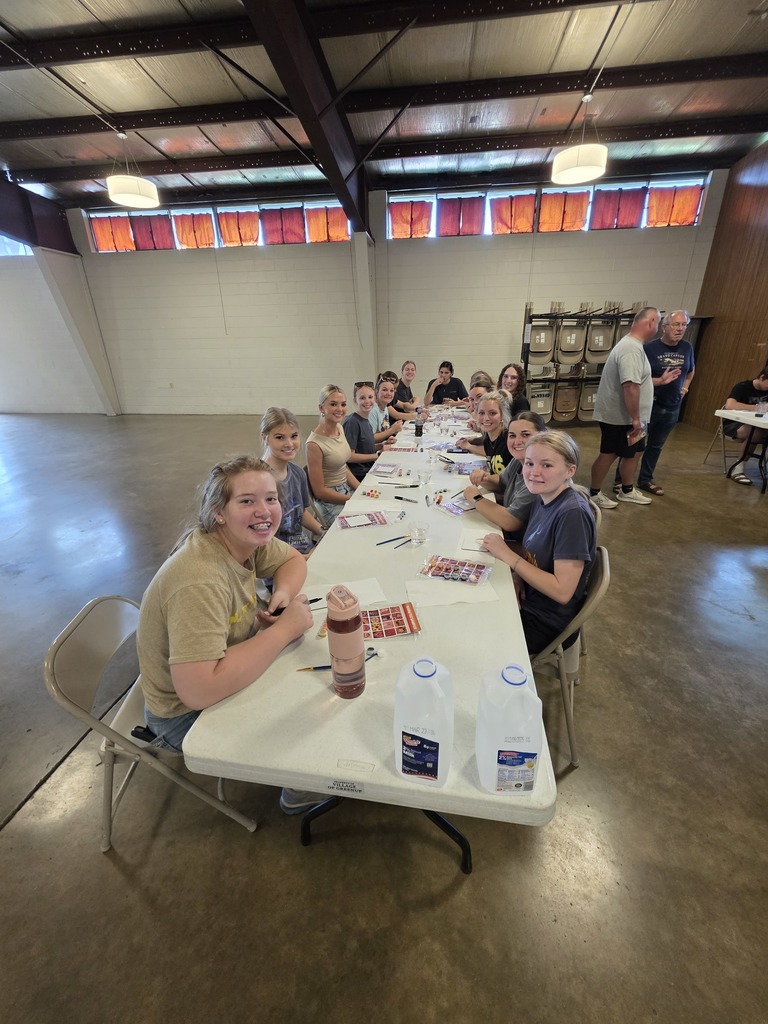 A large group of students are sitting at the tables painting their tiles.