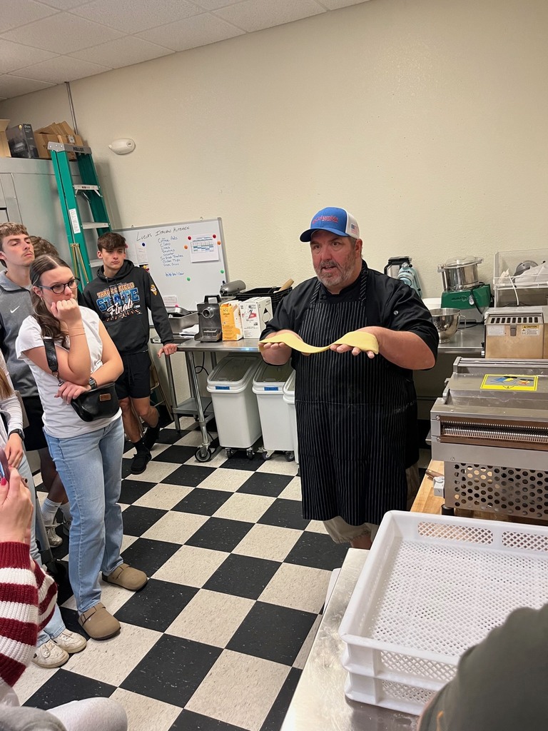 The chef is holding a piece of pasta. Four students are watching him as he speaks. 