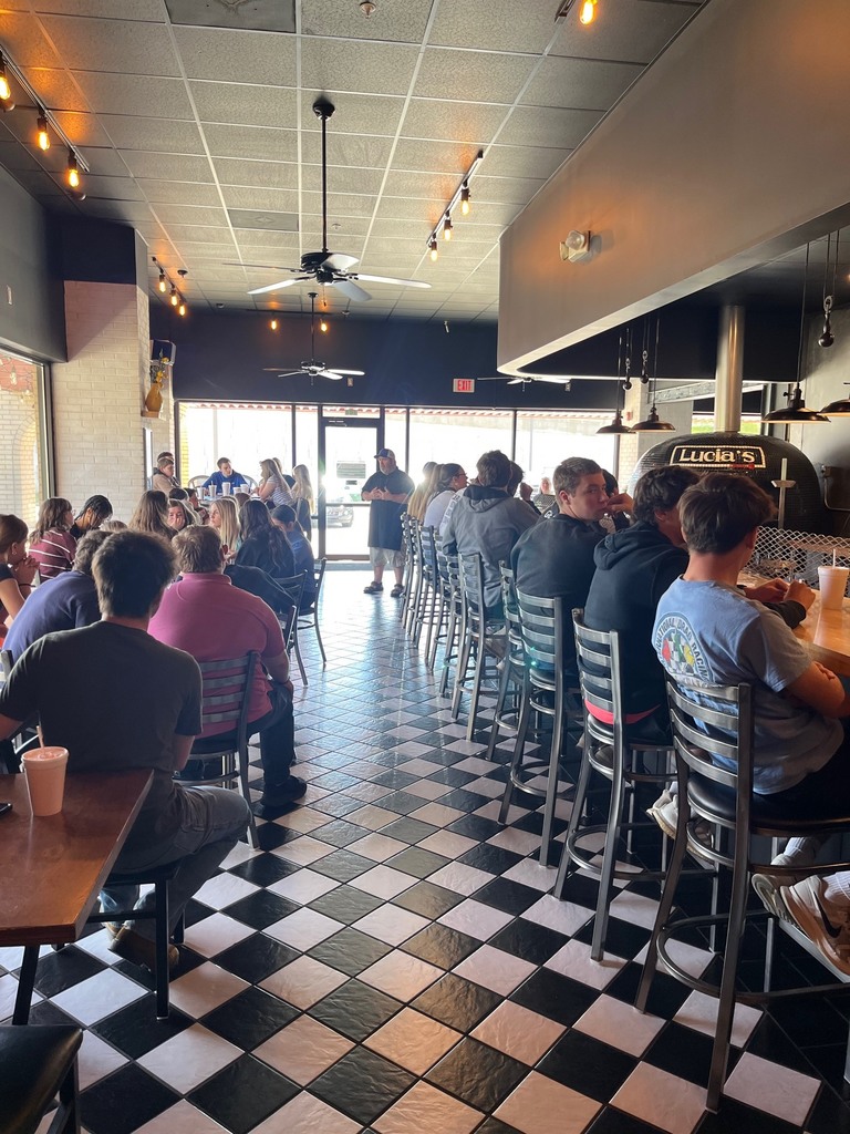 Students are sitting at tables and the bar area. The chef is speaking to the group. 