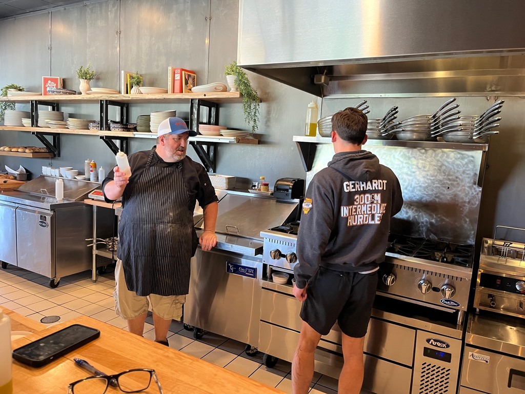 A student is stirring something in a pot and the chef is talking to him. 