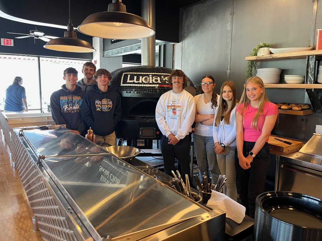 Seven students are standing by the oven in the restaurant. 